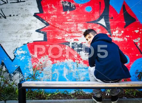 Young man with hoodie sitting on bench in front of graffiti Stock Photos