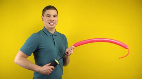 A young man inflates a long red balloon with a pump on a yellow background. Man Stock Footage 143394203