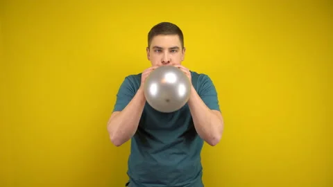 A young man inflates a silver balloon with his mouth on a yellow background. Man Stockbeeldmateriaal 143392687