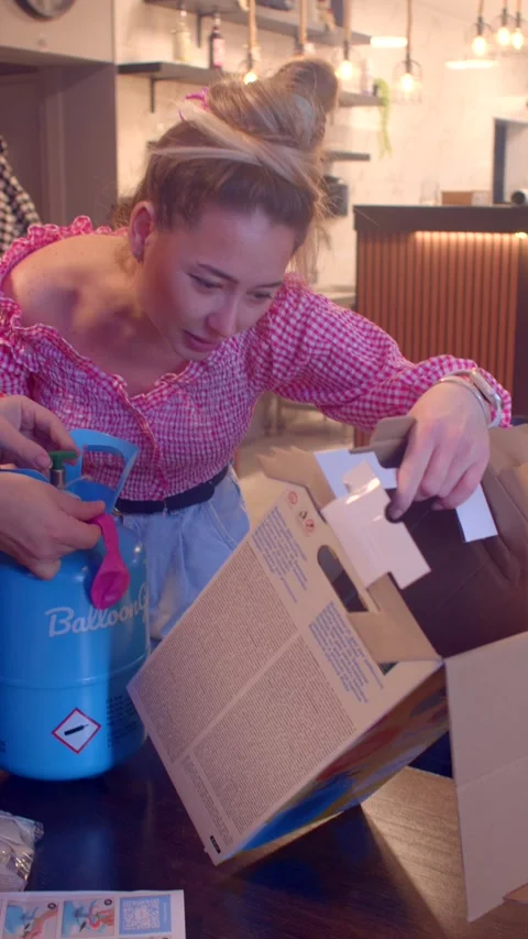 Young man inflating pink balloon with helium tank while woman prepares nearby in Видео 336535489