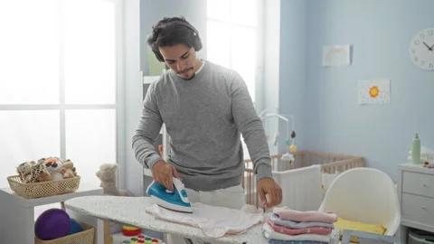 Young man ironing clothes in a cozy bedroom with a baby crib and colorful d.. Stock Footage 306376787