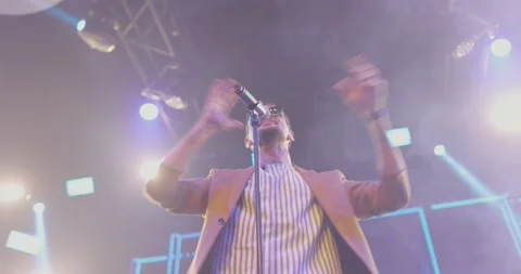 Young man in jacket and black glasses performs song on stage inlight blue Stock Footage 221650254