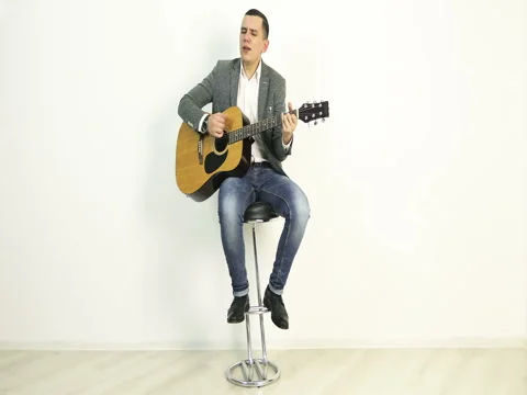A young man in a jacket sits on a bar stool and plays an acoustic guitar. Stock Footage 73778963