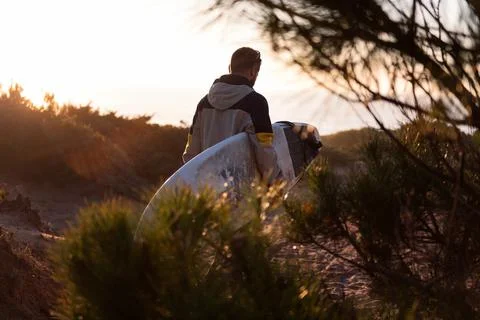 Young man in jacket walking at sunset with his surfboard Stock Photos