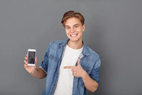 Young man in jeans jacket studio isolated on grey holding smartphone pointing at Stock Photos
