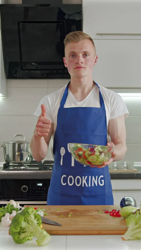 Young man joyfully prepares a fresh salad in a bright, modern kitchen Stock Footage 341761317