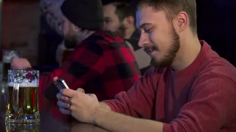 Young man joyfully texting on his smartphone at a lively pub Stock Footage 311986363