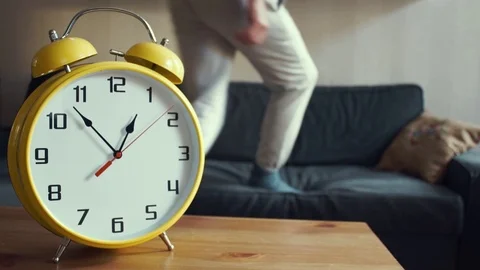 Young man jumping on couch at home. Big yellow clock ticking on the shelf Stock Footage 80814335