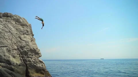 A young man jumping from a high cliff into the sea doing a forward somersault in Video stock 157502270