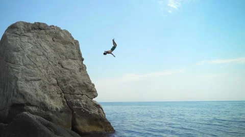 A young man jumping from a high cliff into the sea doing a back somersault in Stock Footage 157502309