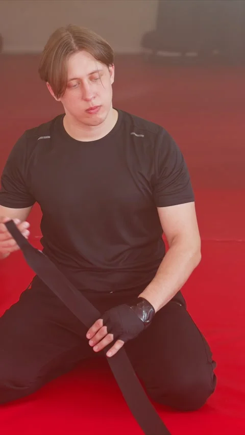 Young man kneeling on red mat preparing and securing black hand wraps for Stock Footage 343081464