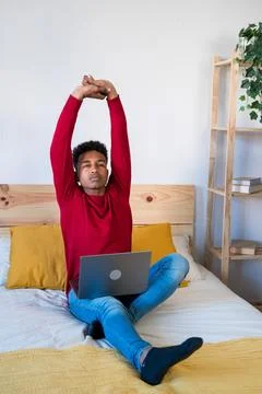 Young man with laptop on bed doing stretching exercises Stock Photos