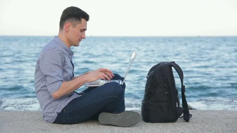 Young man with laptop sitting on the beach typing and browsing the web 库存影片 75970009
