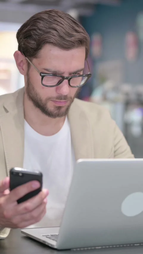 Young Man with Laptop using Smartphone at Work, Vertical Video Stock Footage 342004121