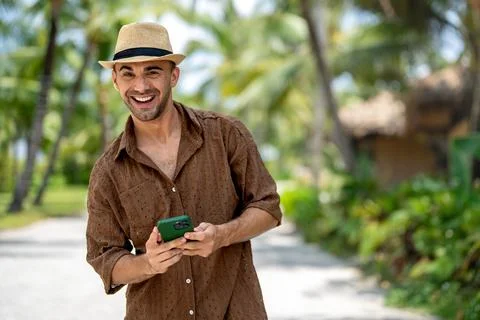 Young man laughing while looking at his smartphone in a sunny tropical reso.. Stock Photos