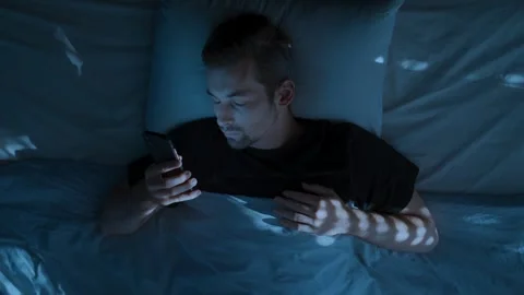 Young man laying in bed at night, holding his smartphone and doom scrolling Stock Footage 314023661