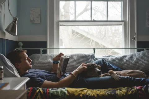 A young man lays on his bed with dog on lap reading by window light Stock Photos