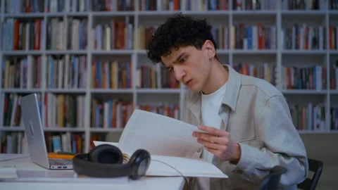 Young Man Leafing Through Book and Studying at Library Table, Laptop and Stock Footage 335123992