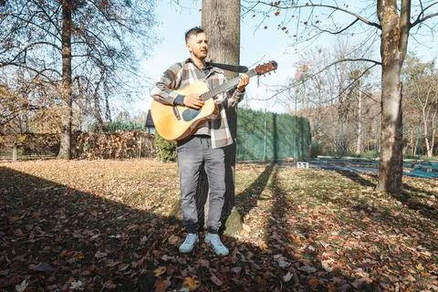 Young man leaning against a tree, playing an acoustic guitar in a sunlit autu 스톡 사진