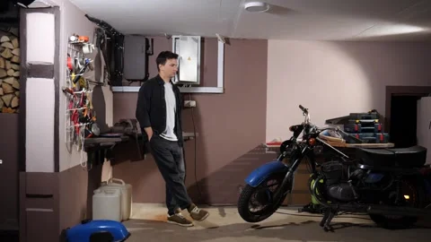Young man leaning against a wall in a home garage, looking at his classic Stock Footage 342758751