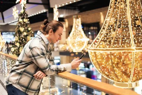 Young man leaning on railing using phone amidst festive Christmas lights an.. Foto stock