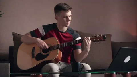 Young man learning to play acoustic guitar online at home Stock Footage 210553516