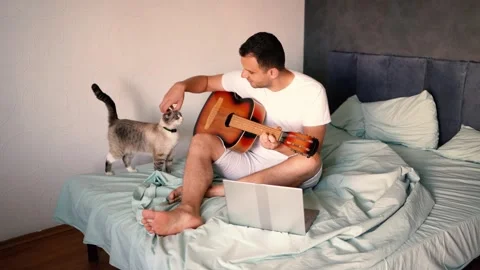 Young man is learning to play the guitar at home, a cat is lying next to hi.. Stock-Footage 276379271