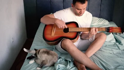 Young man is learning to play the guitar at home, a cat is lying next to hi.. Stock-Footage 276379295
