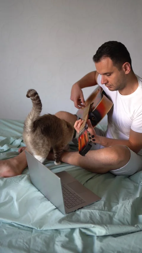 Young man is learning to play the guitar at home, a cat is lying next to hi.. Stock-Footage 276379456