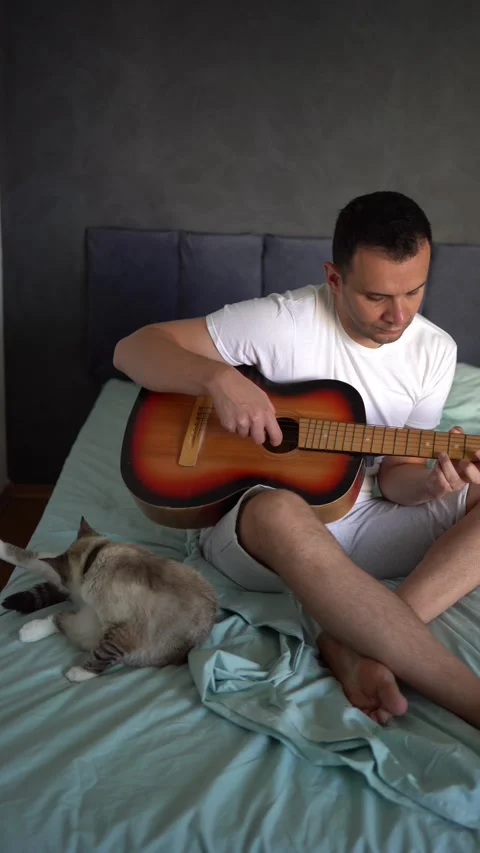 Young man is learning to play the guitar at home, a cat is lying next to hi.. Stock Footage 276379459