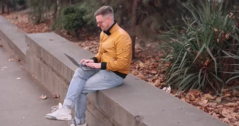 Young man in leather jacket types on laptop sitting on parapet Video stock 308160246
