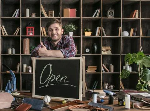 Young man leather maker standing near open sign at table with leather tools Stock Photos