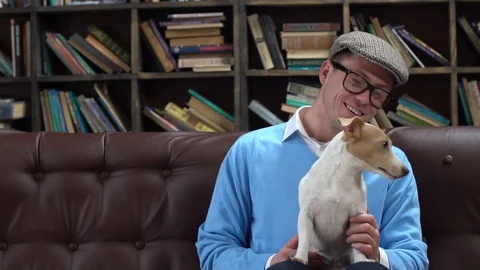 Young man in library in eyeglasses and flat cap sitting holding dog joyful Stock Footage 121539188