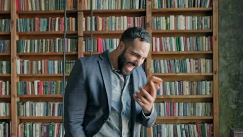 Young Man In The Library With Mobile Phone In His Hands. Something Pleasantly Stock Footage 139909462
