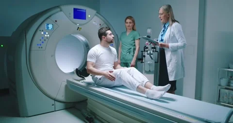 Young man lies down in MRI capsule. Chief head doctor in dressing gown hold Stock Footage 243585347