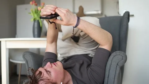 A young man lies upside down in a chair, holding his phone above his head, compl Видео 302634902