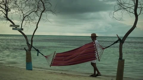 A young man in light clothes approaches a hammock stretched between trees on a Stock Footage 267858180