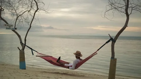 A young man in light clothes lies in a hammock between the trees on the ocean Stock Footage 267847086