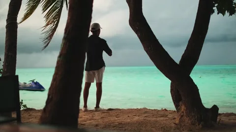 A young man in light clothes stands on a sandy seashore under palm trees. Stock Footage 267852342