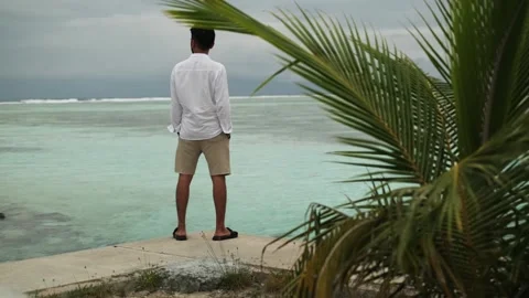 A young man in light clothes walks on a sandy seashore under palm trees. Stock Footage 308088640