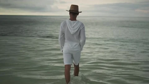 A young man in light white clothes and a hat walks barefoot in the rain on the Stock Footage 310719823