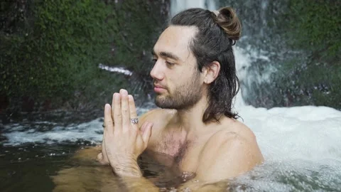 Young man with long haeir sitting in a paryer position in a spring and is Vídeo Stock 198324836