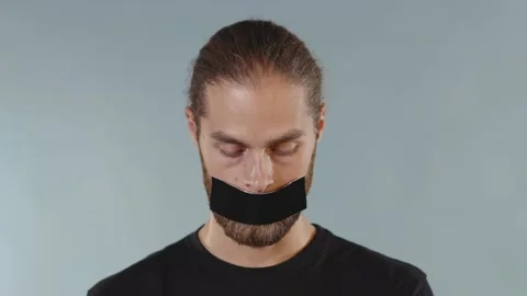 Young man looking at camera serious and tearing tape from her mouth on grey Video stock 133638421