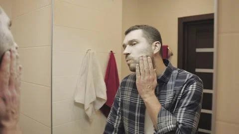 Young man looking in mirror touch his face before shave in bathroom. Slow motion Stock Photos