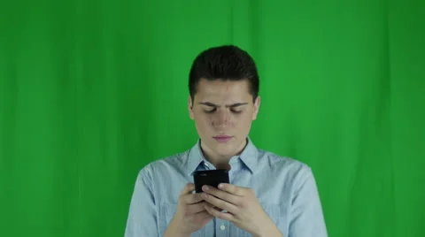 Young man is looking at phone in front of a greenscreen in a blue shirt Vidéo 59083697