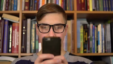 A young man is looking at a smartphone in a library. Man with glasses carefully Stock Footage 121533319