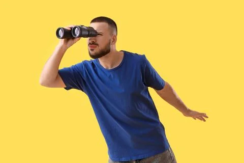 Young man looking through binoculars on yellow background. Search concept Stock Photos