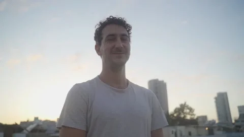 Young man looks at camera and smiles stands on city rooftop at sunset or sunrise Stock Footage 131900828