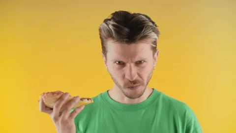 A young man looks into the camera and takes a sharp bite of pizza. A man in a Stock Footage 240925365