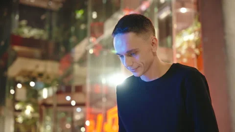 A young man looks down at his laptop ,night street, mid-shot, blurred background Stock Footage 253973218